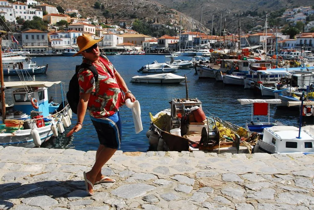 Watercolour artist Scott Pleydell-Pearce, wearing a red hawaiian shirt, sunglasses and sun hat posing in front of Hydra harbour in Greece. 